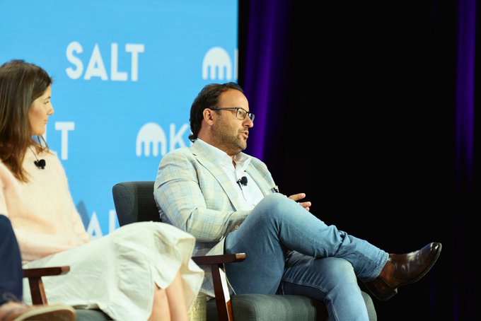 Two panelists, a woman in white and a man in a light blazer and jeans, sit on chairs at the SALT conference with the MK logo visible in the background.