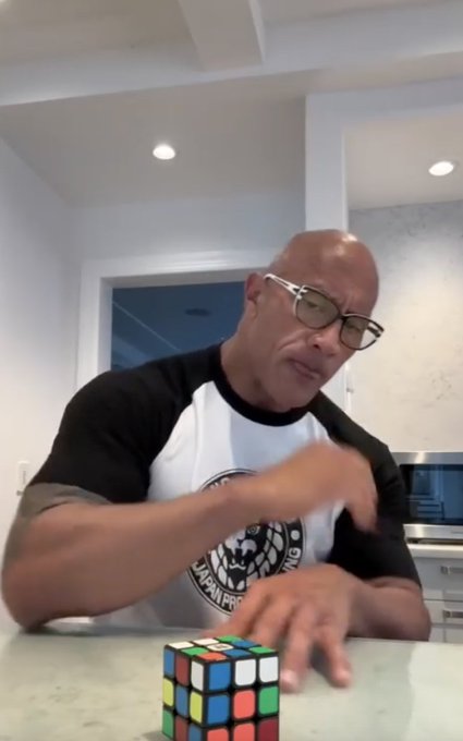 Dwayne "The Rock" Johnson sits at a kitchen counter in a black and white raglan shirt, looking thoughtfully at a solved Rubik's cube in front of him—mirroring the post's theme of trying to solve the complex puzzle of sustainable crypto yields and warning that anything too convoluted to explain simply (like the colorful cube's many moves) likely disguises emissions or unsustainable loops.