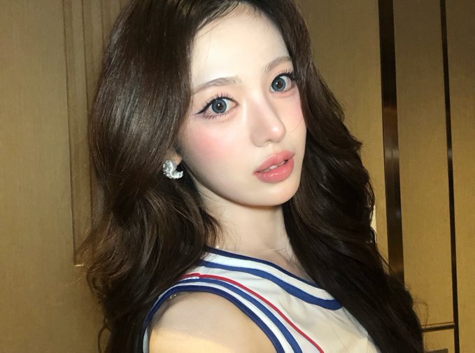 A close-up selfie-style photograph of a young woman with long dark wavy hair, wearing a striped top and earrings, looking at the camera.