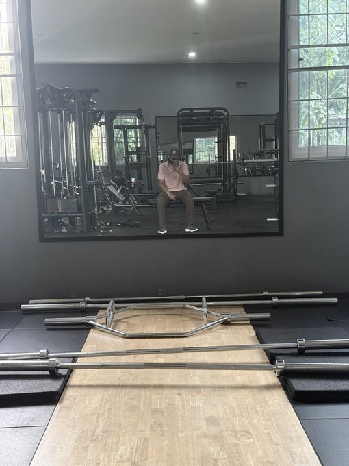 A man in a pink shirt sits on a weight bench inside a gym, taking a photo in a mirror, with a hexagonal trap bar and barbells resting on the wooden lifting platform in the foreground.