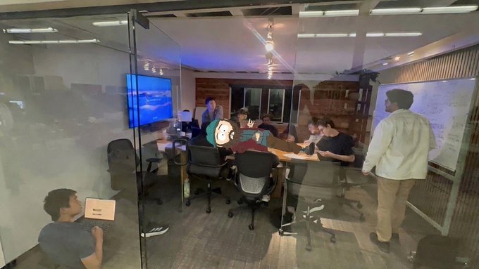 A group of people gather around a conference table in a glass-walled meeting room for what appears to be a collaborative work session or meeting, with a wall-mounted TV displaying an image, whiteboards visible, and one person standing to address the seated team.