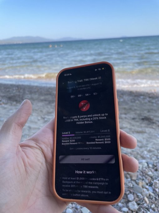 A hand holds an orange smartphone displaying a 'Trading Trail: TRX (Week 2)' campaign page with a countdown timer and reward details, set against the backdrop of a beach and ocean.