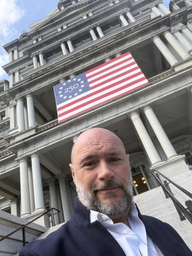 A bearded man in a dark suit takes a selfie in front of a grand neoclassical building, likely a U.S. government structure, with a large American flag featuring the original 13-star "Betsy Ross" design hanging prominently from the facade. The "Chiliz goes stateside" caption suggests this marks the blockchain sports platform's expansion into the American market, with the patriotic backdrop symbolizing their entry into the United States.