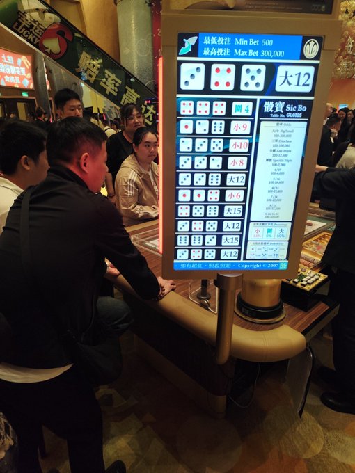 People are gathered around a brightly lit Sic Bo gaming table in a casino, with a large display screen showing the betting options, dice combinations, and payout amounts.