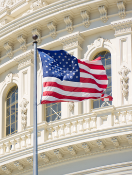 An American flag waves in the foreground against the ornate white facade of a classical government building. The image symbolizes federal authority, providing a visual backdrop to the news of personnel turnover within the White House Crypto Council, suggesting instability at the intersection of government and cryptocurrency policy.
