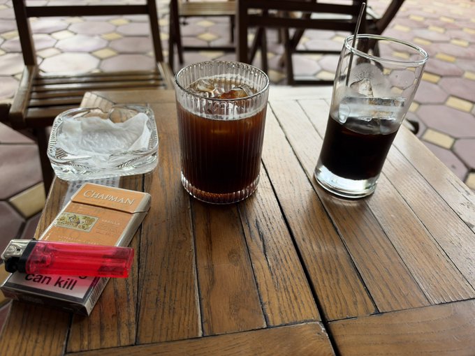 A wooden café table holding two glasses of iced coffee, a glass ashtray, a red lighter, and a pack of cigarettes with the visible text 'can kill'.