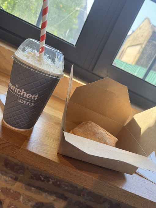 A takeaway coffee from "Enriched Coffee" with a red and white striped straw sits beside a pastry in a cardboard box on a wooden windowsill. The image visually supports the post's offer to buy a coffee, reinforcing the "Gm" greeting and the promotional call-to-action for followers to engage.