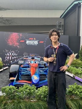A young man wearing glasses and a lanyard stands next to a blue and red Formula 1 car on display, with a "Singapore Grand Prix" backdrop visible behind him. He flashes a peace sign in what appears to be a paddock club area, reflecting the post's celebration of fulfilling his mechanical engineering dream by attending the F1 Singapore Grand Prix.

**Visible text:** "SINGAPORE GRAND PRIX" on the backdrop.