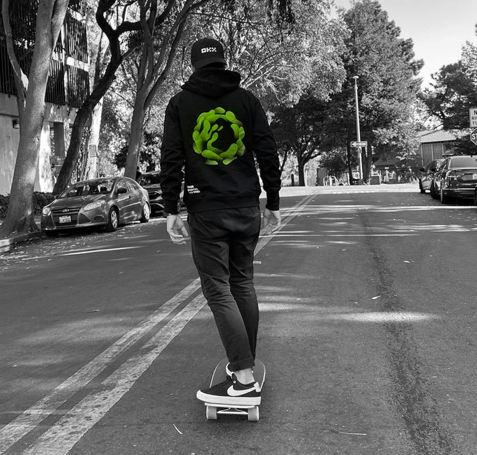 A skateboarder rides down a tree-lined street wearing a black cap marked "DHX" and a black hoodie featuring a bright green spiral logo, paired with Nike sneakers, perfectly capturing the laid-back vibe of "rolling into the weekend."