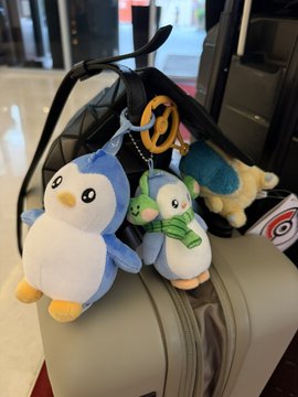 A set of plush keychains featuring penguins and another character hanging from a black bag placed on top of a suitcase.