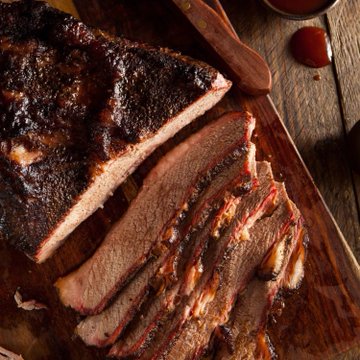 Sliced smoked brisket with a dark, seasoned crust is arranged on a wooden cutting board, with a small bowl of sauce and wooden utensils nearby.