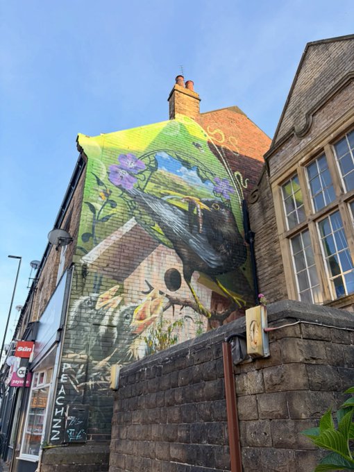 A vibrant street mural on a brick building depicting a large black bird alongside flowers and natural scenery, photographed from below against a clear blue sky.
