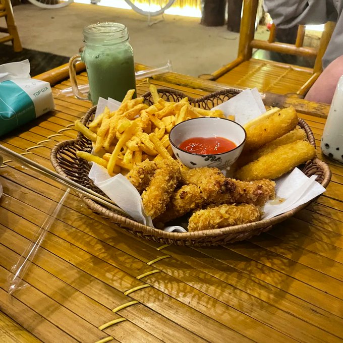 A basket containing french fries, fried chicken pieces, and fried mozzarella sticks with a side of ketchup and a green iced drink, served on a bamboo table.