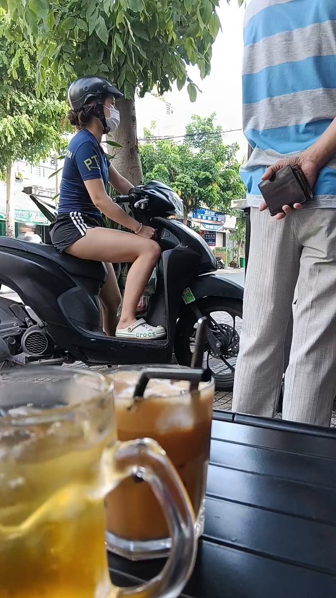 The image shows a man wearing a blue shirt sitting on a motorcycle, while another person stands nearby with their handbag. There are two cups of tea placed next to them, suggesting they might be enjoying a break or having a conversation over some refreshments. The scene takes place in an outdoor cafe setting, and the post about the cafe is titled "Nay có lương paid khá ổn rồi Vợ nói ngồi uống cafe rồi cày đi Còn đân sướng A.E nhỉ".