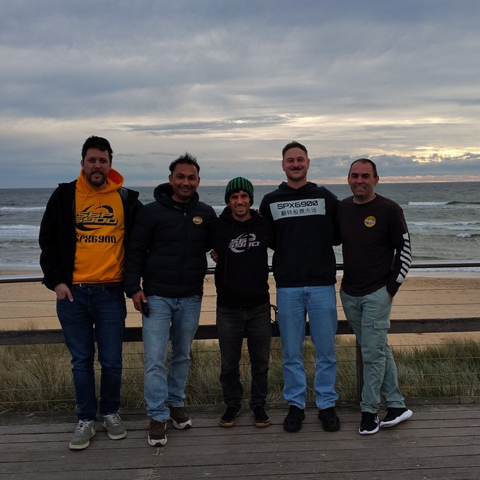 Five members of the SPX6900 community pose together on a wooden boardwalk at a beach in Melbourne at sunset, with the ocean and a cloudy sky in the background. Two of the hoodies and one shirt prominently display the "SPX6900" logo, matching the post about a SPX6900 meetup gathering of "Aeons" in Melbourne 2026.