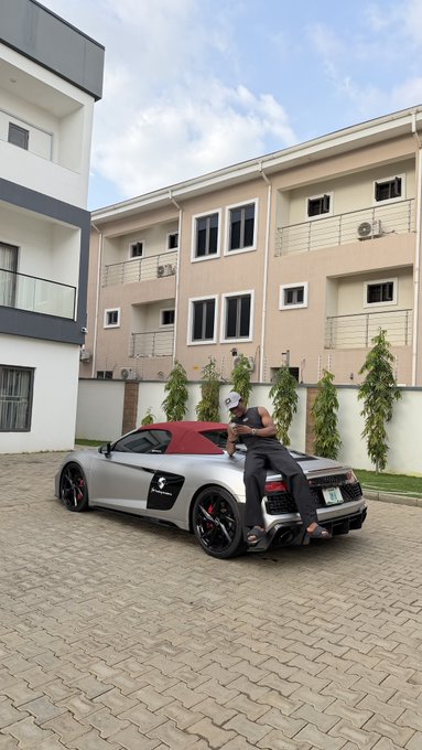 A matte silver Audi R8 Spyder with a red soft top and orange brake calipers is parked in a paved driveway in front of a modern two-story duplex, where a man in a black shirt stands beside it appearing to interact with someone inside.

The image humorously illustrates the post's "Crypto bro vs Forex bro" showdown theme, with the luxury sports car serving as a symbol of the flashy lifestyle rivalry between the two trading communities being joked about.