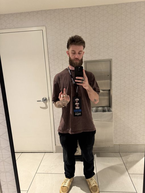A man takes a mirror selfie in a restroom, wearing a brown t-shirt with a lanyard displaying "MOON" text, and throwing up a peace sign in response to the "GM" greeting request.
