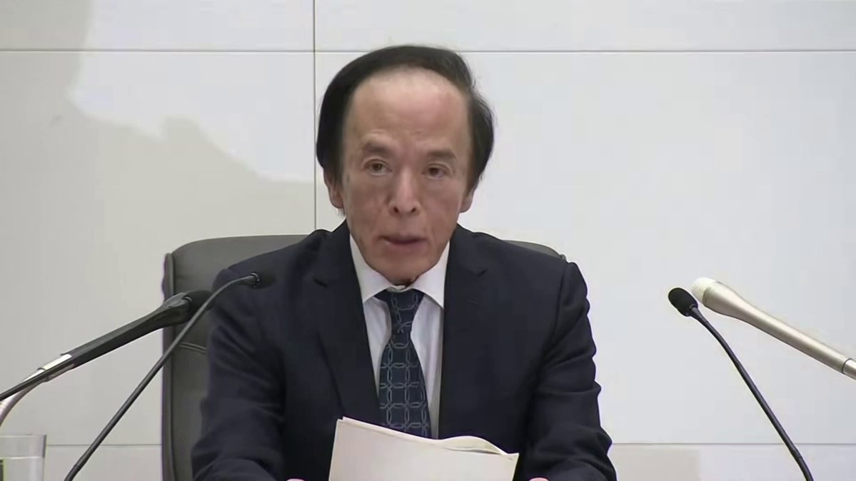 The image shows a man dressed professionally sitting at a desk with two microphones, presumably for a press conference or interview. The man appears to be giving a speech or announcement about new interest rates that are expected to be announced soon. The context suggests that the event is related to the Japanese economy and financial markets, as it is referred to as a "breaking news" event based on market speculation. This scenario implies that markets are pricing in an unusually high probability for the BOJ to hike interest rates to 100 BPS.
