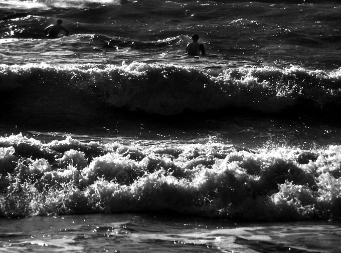 A black-and-white photograph captures two silhouetted figures in the ocean as large, foamy waves roll toward the shore.