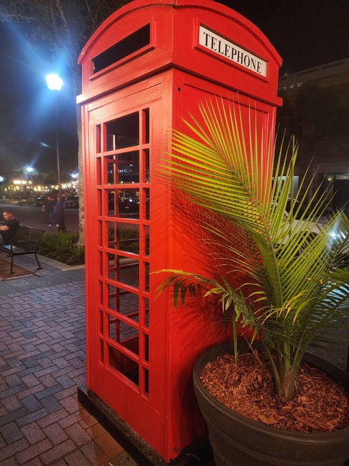 A classic red British telephone booth standing on a brick sidewalk at night, next to a potted palm plant, with a street lamp and people visible in the background.