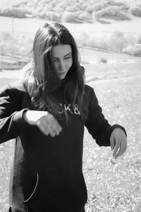 A black-and-white outdoor portrait of a young woman with long hair wearing a black hoodie with partial lettering visible, looking downward while standing in a grassy field.