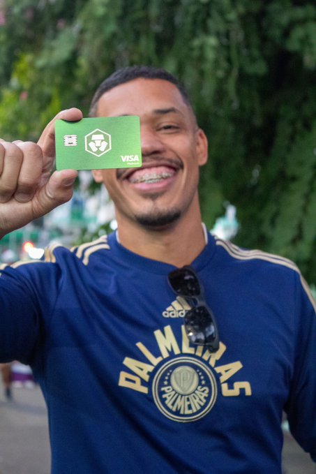 The image features a man wearing a blue shirt with the word "Palmeras" written on it. He is holding up a green Visa card, which has a palm tree design on it. The man appears to be smiling for the camera while showing off his card.