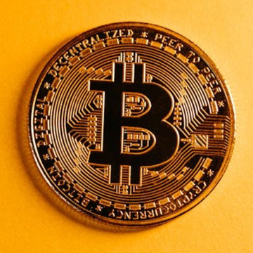 A gold-colored physical Bitcoin token rests on a yellow surface, displaying the '₿' logo and the text 'DECENTRALIZED PEER TO PEER' and 'BITCOIN DIGITAL'.
