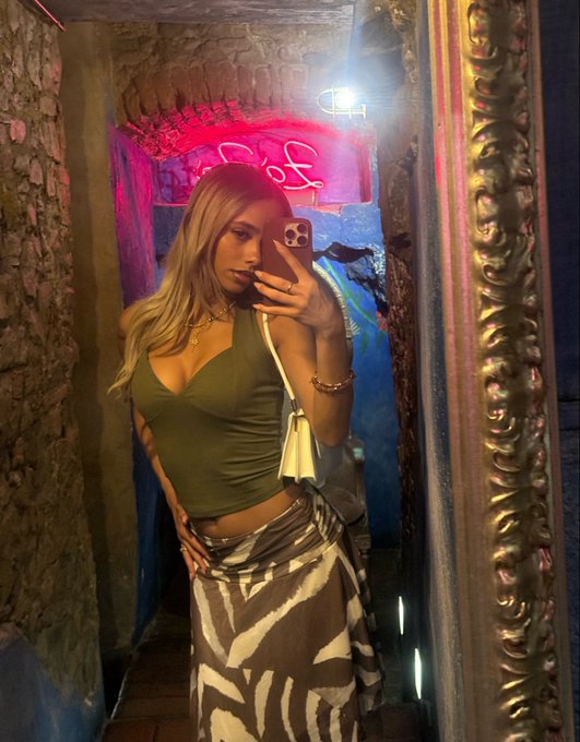 A mirror selfie of a blonde woman wearing an olive green top and a zebra-print skirt, standing in a dimly lit venue with neon signage in the background.