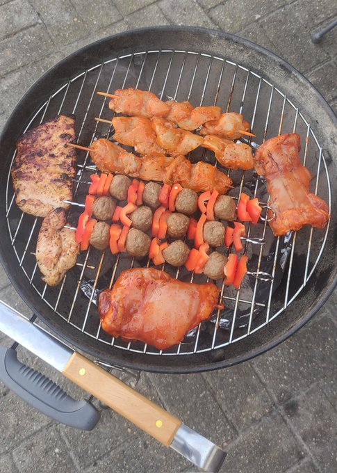 The image shows a variety of food items, including meat and vegetables, being cooked on an outdoor grill. The grill has several skewers with different types of food, such as chicken wings, beef, and carrots. A metal spatula can be seen resting on top of the grill, ready to flip or turn the food. The scene is set outdoors in a backyard, suggesting that it might be a casual gathering or a family meal.