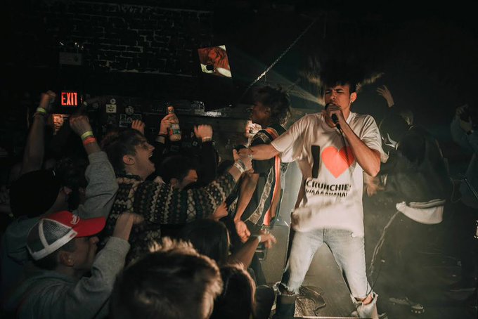 A performer in a white Crunchie t-shirt holds a microphone while singing energetically on an energetic concert stage, with audience members reaching toward him.