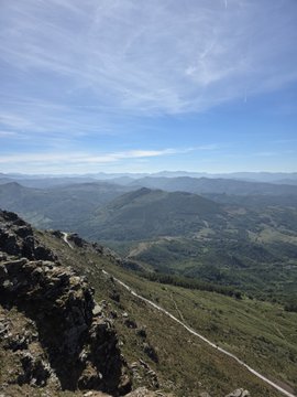 A scenic mountain landscape viewed from a rocky peak, showing a winding trail descending into rolling green hills under a bright blue sky with wispy clouds.