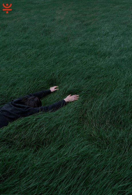 An atmospheric photograph of a person lying in tall, dark green grass with arms outstretched, featuring a small red logo in the top left corner.