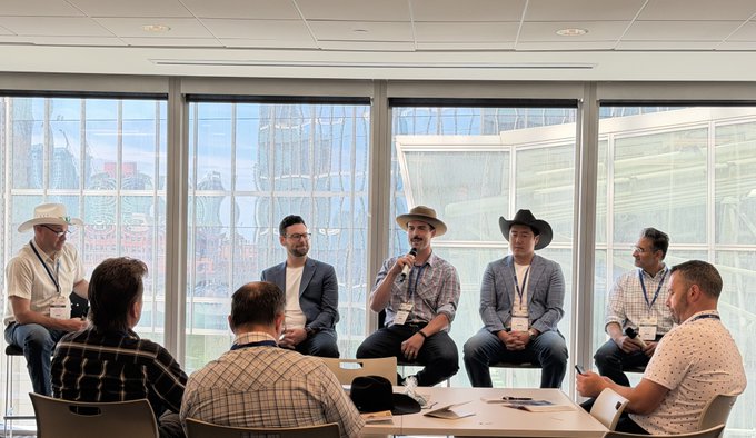 A panel discussion featuring several men, some wearing cowboy hats, seated in a room with large windows overlooking a city, with one man holding a microphone while others listen.