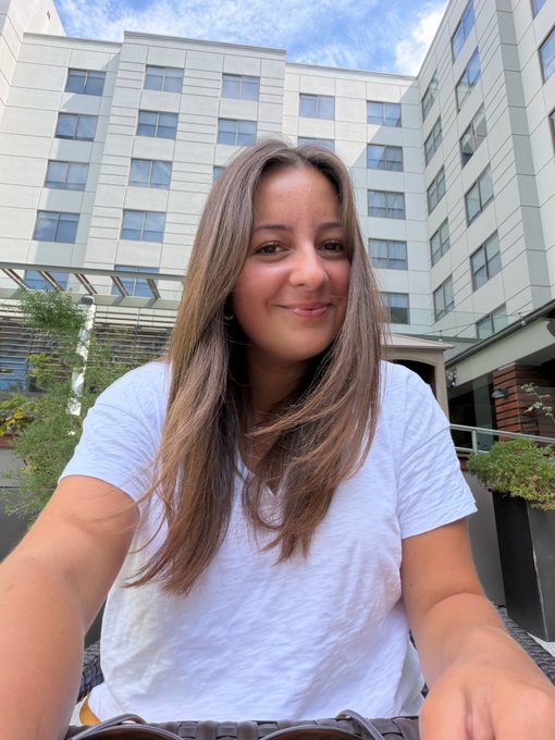 A young woman with long brown hair takes a selfie wearing a white t-shirt outdoors with a modern building behind her.