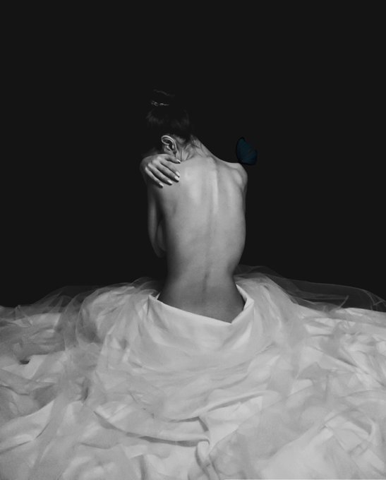 A black and white artistic photograph of a woman's bare back as she sits on the floor surrounded by white fabric, with a single blue butterfly resting on her shoulder.