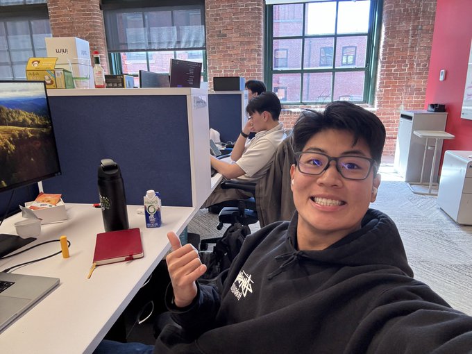 A young man in a black hoodie smiles and gives a thumbs-up at his desk in a modern office with brick walls, with "ULUI" boxes and a laptop visible nearby, capturing the excited "day one" moment of his $10,000 AI Polymarket trading bot project.