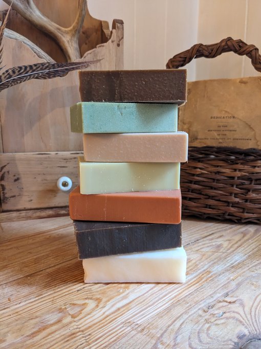 A stack of seven colorful artisan soap bars in shades of brown, mint, peach, green, terracotta, dark brown, and cream, displayed on a wooden surface next to a basket and decorative antlers.