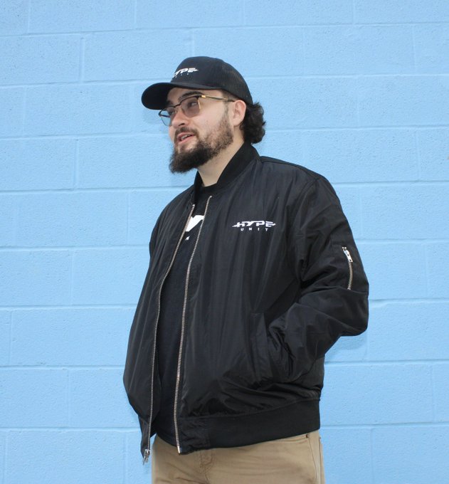 A man with a beard and glasses wearing a black 'HYPE' cap and matching black bomber jacket stands in front of a light blue cinder block wall.