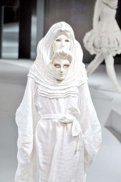 A surreal white sculptural figure or mannequin featuring multiple stacked masked faces beneath a draped hood, displayed in a gallery setting with another similar white sculpture visible in the background.