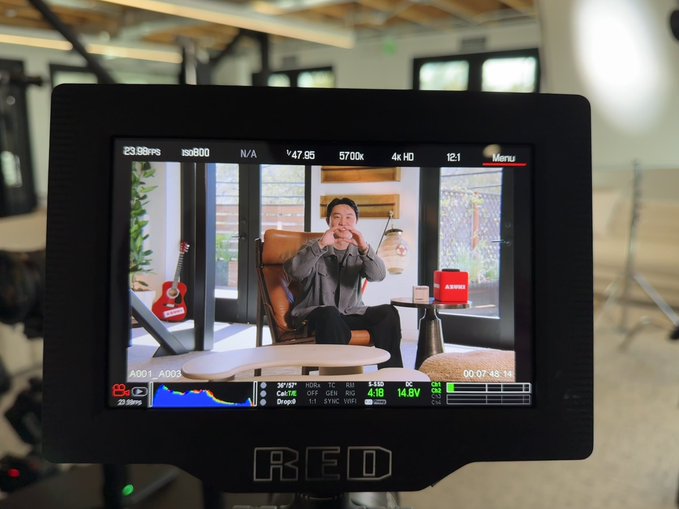 A RED camera monitor displays a live feed of Zagabond, co-founder and CEO of Azuki, seated in a brown chair with a red guitar and Azuki-branded box in the background. The monitor shows recording settings including "23.98p," "ISO800," "4K HD," "5700k," and a runtime of "00:07:48:15," indicating an interview or documentary production about the Azuki NFT founder.