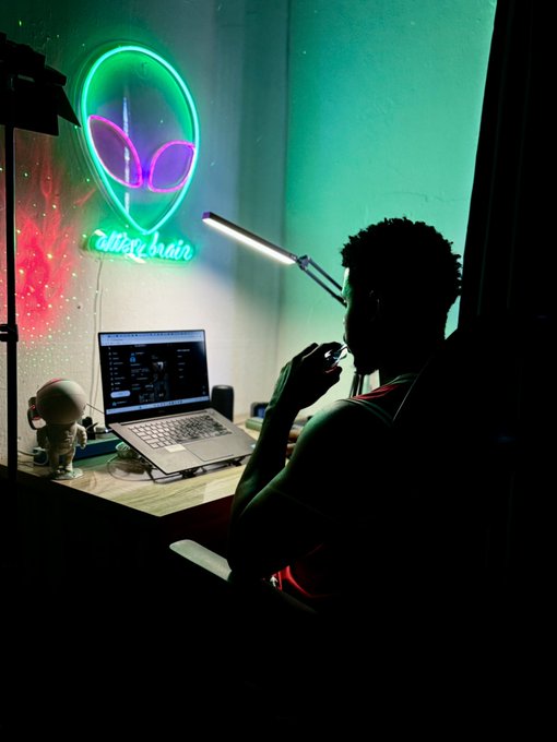 A silhouetted person sits at a desk working on a laptop in a dimly lit room illuminated by a green alien-themed neon sign and a desk lamp.