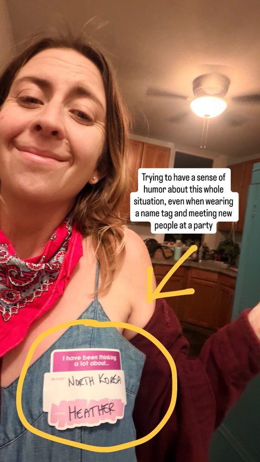 A selfie of a smiling woman named Heather wearing a name tag that humorously lists "North Korea" as her country, with a caption explaining she's trying to maintain a sense of humor about wearing the name tag at a party.