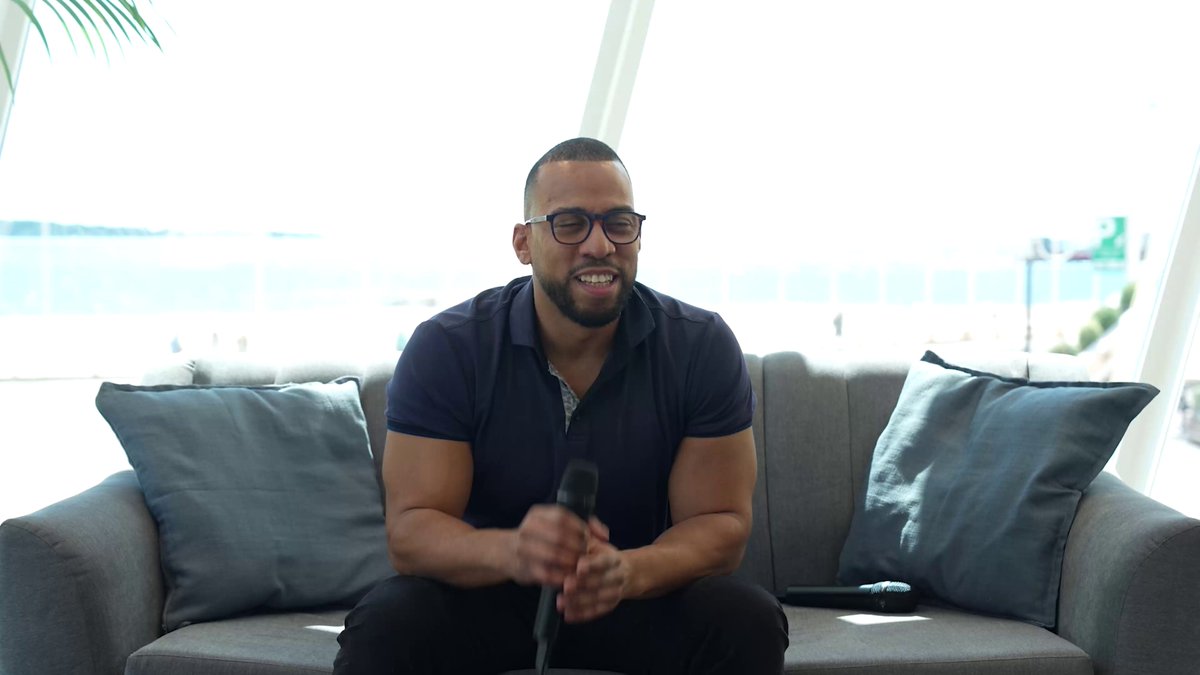 The image features a man sitting comfortably on a couch, holding a microphone. He appears to be smiling as he looks directly at the camera. The setting seems to be indoors, possibly in an office or a studio environment. There are two blue pillows placed behind him and another pillow further back on the couch.