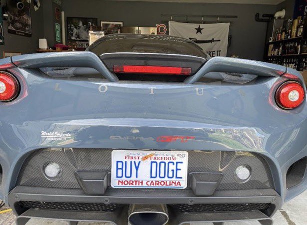 A light blue Lotus Evora GT410 sports car is shown from the rear in a garage, featuring a custom North Carolina license plate reading "BUY DOGE," which playfully ties into the accompanying post's message about "First in Freedom" and supporting Dogecoin ($DOGE).