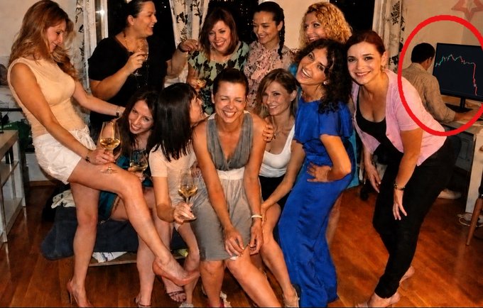 A group of women posing together at an indoor party, with a red circle highlighting a man in the background facing away near a computer monitor displaying a chart.