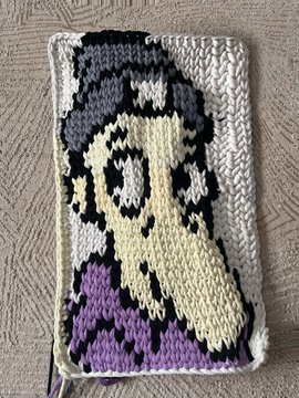 A handmade crochet or knit piece depicting a stylized female character with long hair in a purple dress, displayed on a textured fabric background.