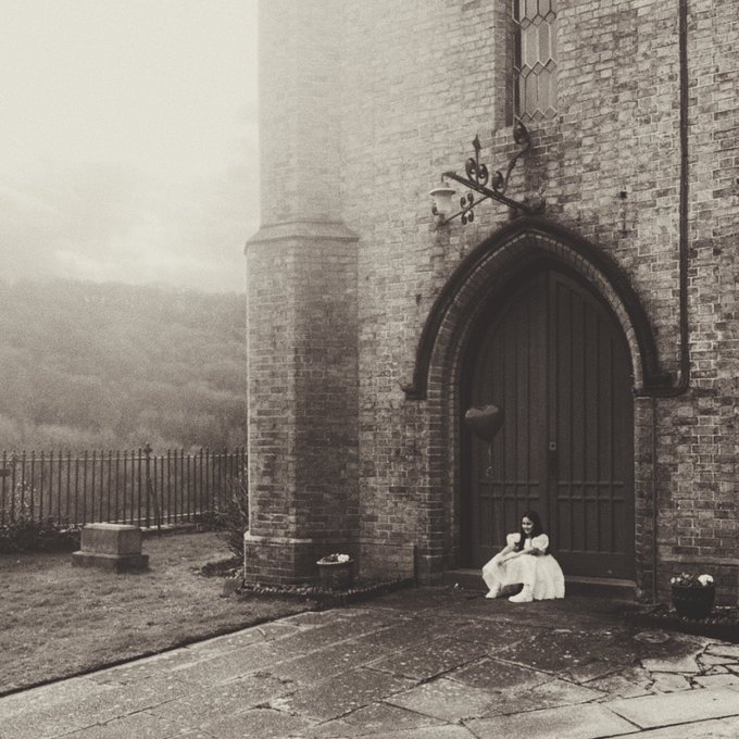 A sepia-toned photograph shows a young girl in a light dress sitting on the ground in front of the large wooden doors of a stone church, with misty hills visible in the background.