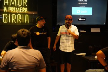 Two men presenting at an event, with a neon sign reading 'AMOR A PRIMERA BIRRIA' and a screen displaying 'Gracias' and '1.1 alpha'.