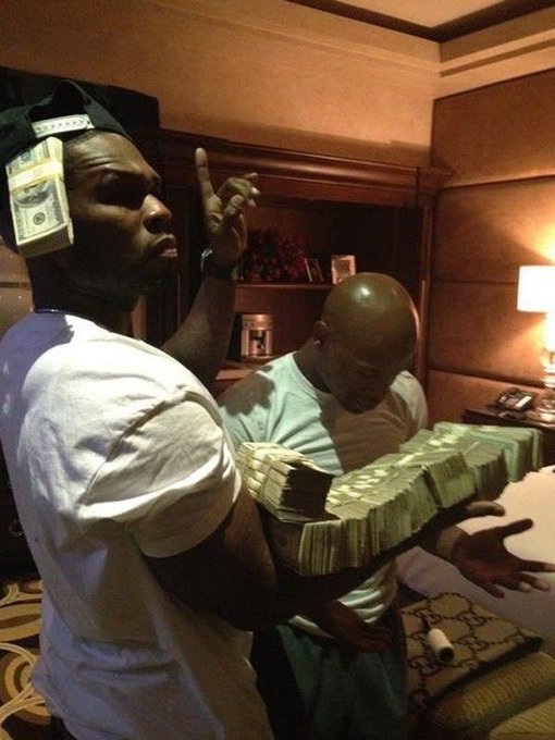 Two men pose in a hotel room with thick stacks of cash, one wearing a black cap with bills stuffed in the strap and another holding a massive bundle of banded money. The image complements the post's confident $120 stock call by projecting an image of wealth and big wins.