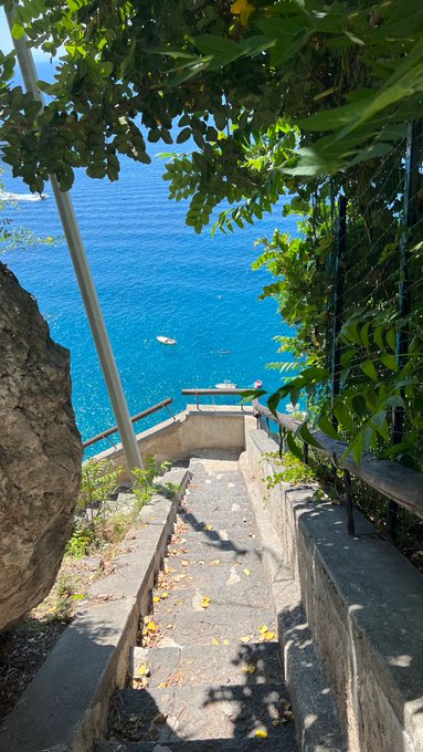The image shows a stone staircase leading down towards the ocean. There are two boats visible near the water's edge. The sky above appears hazy, creating an atmospheric setting for this picturesque scene.